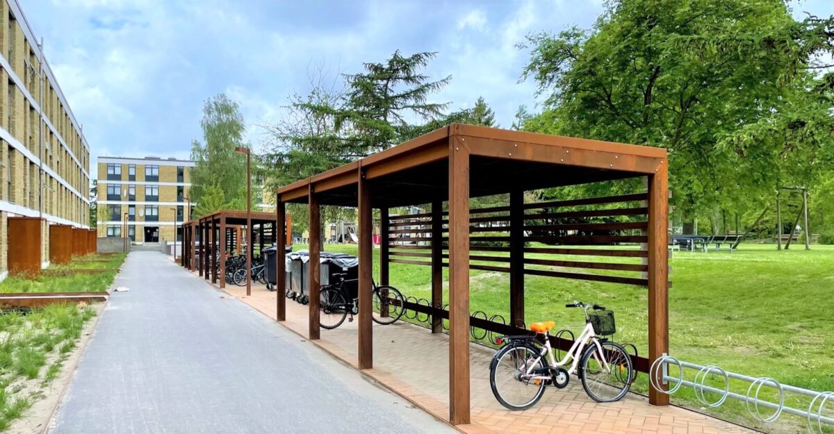 Casa 44 en modulopbygget cykeloverdækning i cortenstål med installeret cykelstativer og beklædning af mahogni planker, som står ved siden af en stor lejlighedsbygning i Vollsmose